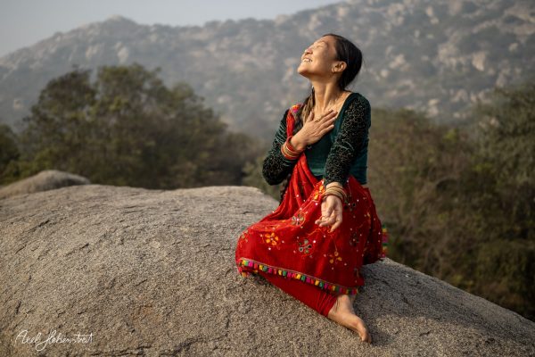 Woman in traditional attire expresses joy while sitting on a rock, surrounded by nature and mountains.