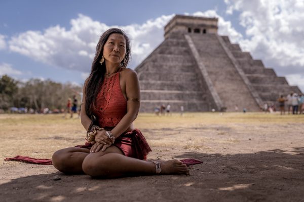 A woman in a red dress sits gracefully in front of the ancient pyramid at Chichén Itzá, exuding tranquility.