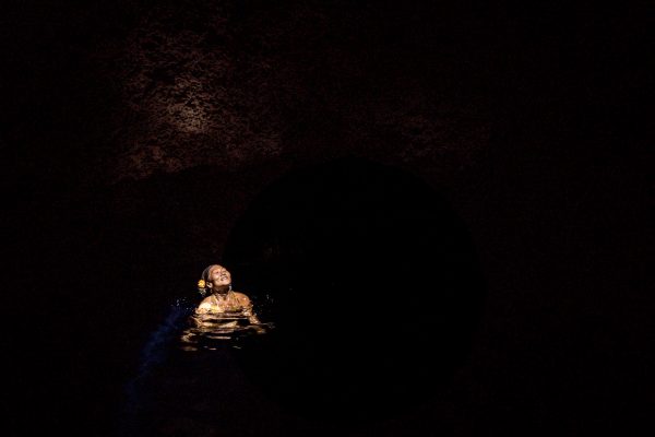 A woman in a golden outfit gracefully floats in dark waters, illuminated softly by surrounding cave walls.