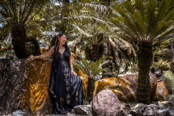 Woman in a black dress poses serenely among lush greenery and palm trees, creating a tranquil, exotic atmosphere.