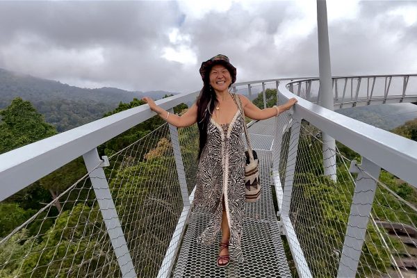 A woman in a patterned dress stands joyfully on a suspension bridge surrounded by lush mountains and fog.