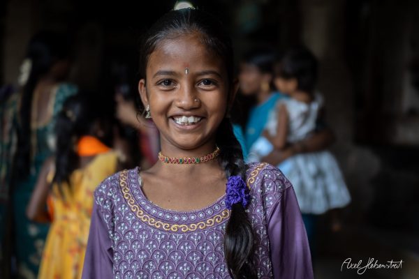 Smiling girl in traditional attire with a purple dress and choker, surrounded by children in a festive atmosphere.