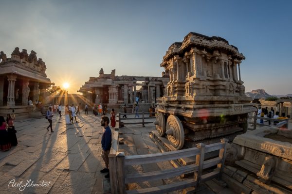 Sunset casts warm light over ancient stone architecture in Hampi, bustling with visitors exploring the historic site.