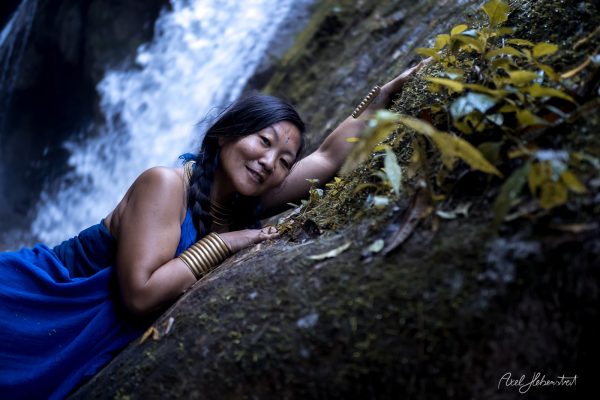 A woman in a blue dress lies peacefully on a moss-covered rock by a waterfall, surrounded by nature's greenery.