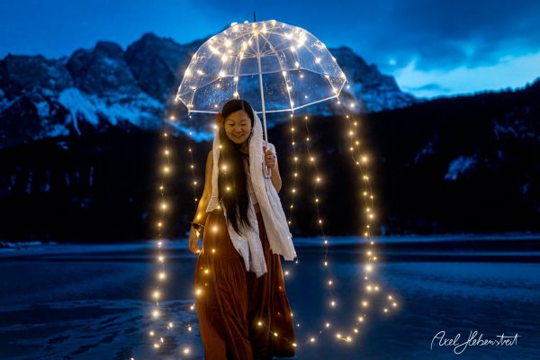 Woman in a flowing dress smiles under a light-up umbrella by a mountain lake at dusk, creating a magical atmosphere.