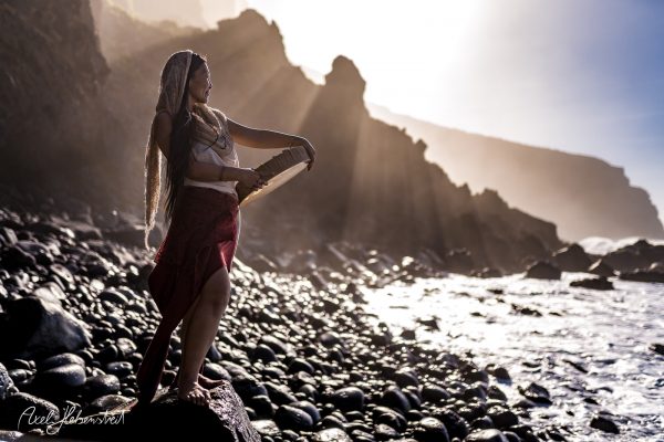 A woman with long hair plays a musical instrument on a rocky beach, illuminated by sunlight and misty cliffs.