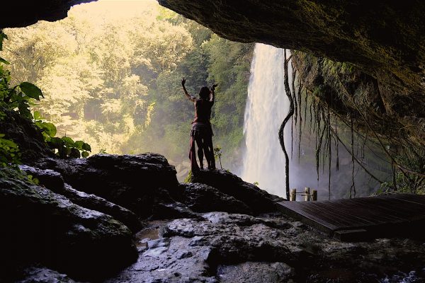 A person stands triumphantly in a cave, overlooking a waterfall surrounded by lush greenery.