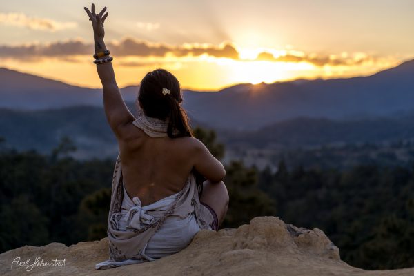 A person with long hair sits on a rocky outcrop, raising their arm towards a sunset over distant mountains.