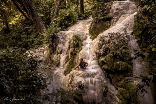 A person sits on moss-covered rocks beside cascading water in a lush, tropical forest setting.