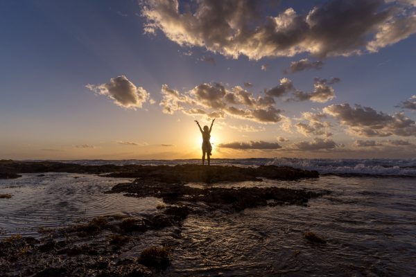 Person celebrating with arms raised on rocky beach at sunset, with waves and clouds creating a dramatic atmosphere.