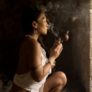 A woman performing a spiritual ritual with smoke in a serene, dimly lit stone setting.